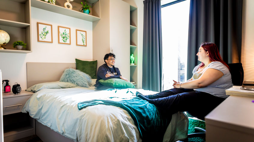Two people in a university halls room. One is on the bed, the other is at the desk.