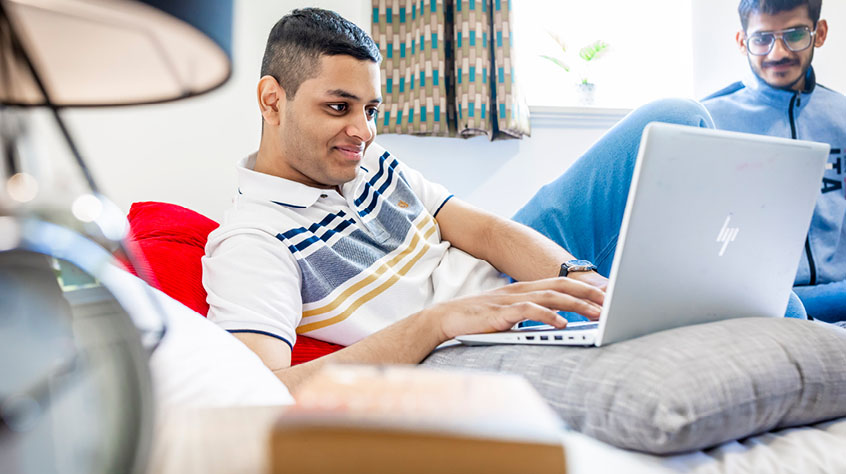 Two students in a communal space, one of the students is working on a laptop