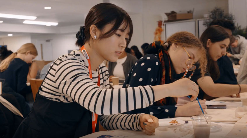A group of individuals seated at tables, each holding paintbrushes, engaged in a painting activity.