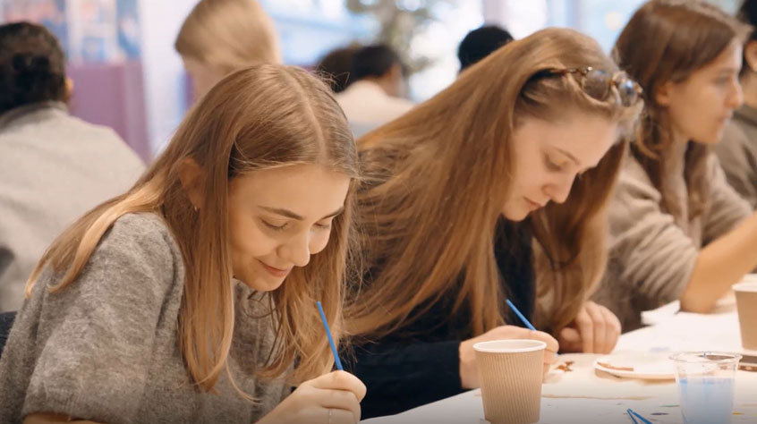  A group of young individuals gathered around a table, engaged in painting with paint and a paint brush.